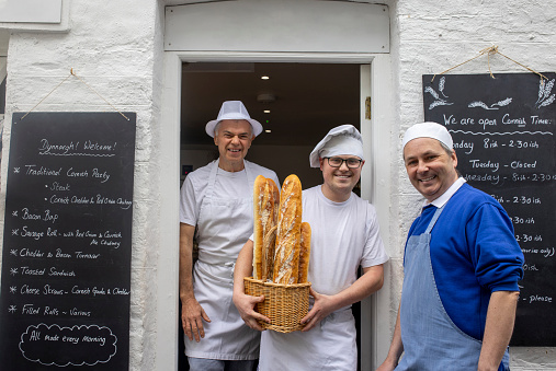 Bakery workers standing outside of their small bakery business in Polperro, Cornwall. They are smiling looking at the camera and the man in the middle is holding a basket of freshly baked baguettes.