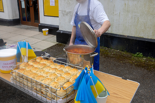 Medium shot of homemade Ukrainian garlic bread rolls and borscht soup being sold for charity in Polperro, Cornwall. An unrecognisable man is setting up the event wearing an apron. There are Ukrainian mini flags on the table.
