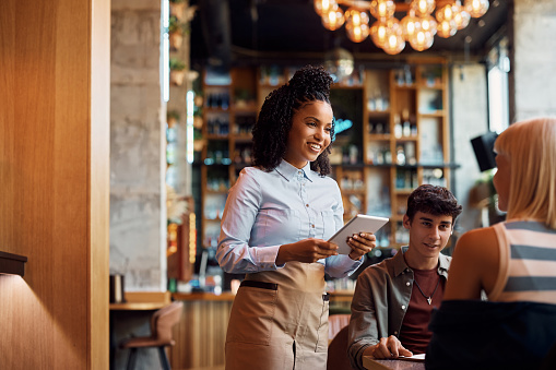 Happy African American waitress using touchpad while taking order from customers in a restaurant.