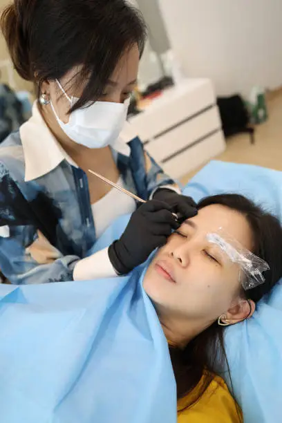 An eyebrow embroiderer is helping a woman embroider eyebrows in her salon studio.