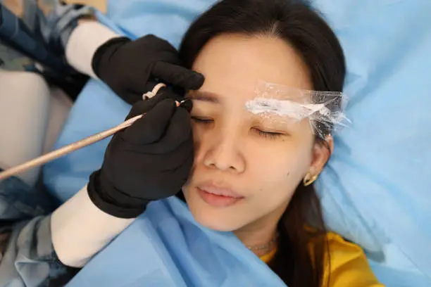 An eyebrow embroiderer is helping a woman embroider eyebrows in her salon studio.