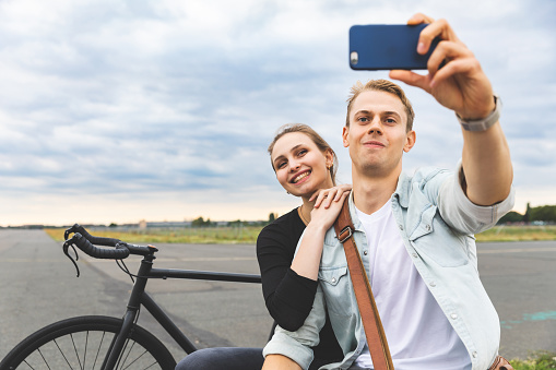 Happy couple together taking a selfie and smiling - Man and woman sharing time together, sitting next to a fixed gear bike and looking at the phone - friendship and lifestyle concepts