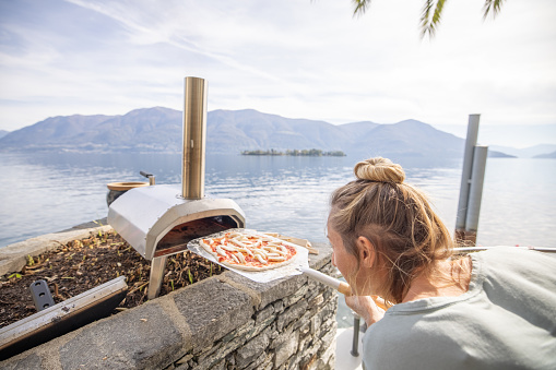 Making Italian pizza at home in the garden using a small domestic wood fire pizza oven, baking fresh pizza with a stunning view of lake Maggiore