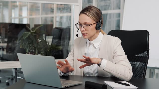 Female executive wears headset video calling on laptop. Businesswoman webinar speaker streaming live web training. Call center agent, service support manager speaking to distance customer in webcam.