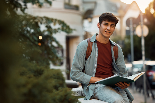 Smiling teenage boy seated on the concrete stairs outside while perusing a textbook in his hands