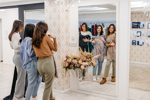 Three beautiful young female friends shopping together for prom dresses in the boutique. Looking in the boutique mirror.