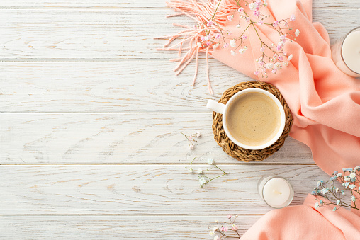 Hello spring concept. Top view photo of cup of fresh coffee on rattan serving mat candles in glasses gypsophila flowers and pink soft plaid on grey wooden table background with copyspace