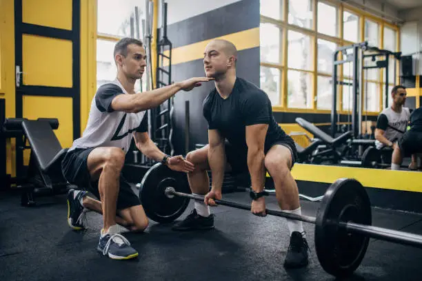 Two men, fit male exercising with weights, while his personal trainer is assisting him in gym.