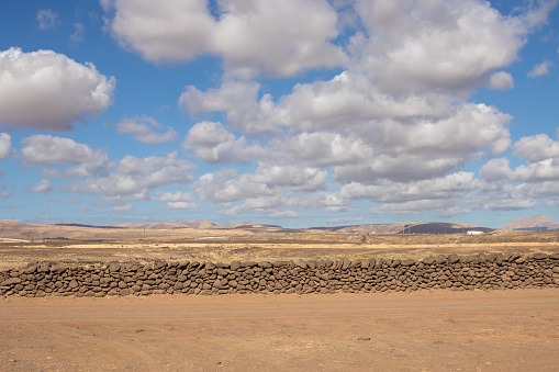 Landscape with an orange soil in the sunshine. Mountains in the background. Stone fence in the foreground. Blue sky with white clouds. Puerto Lajas, Fuerteventura, Canary islands, Spain.