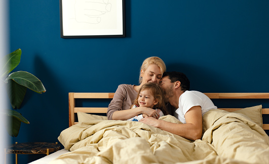A loving couple lying in bed in the morning and cuddling with their young daughter. Mother is hugging the girl while the father is leaning on her shoulder. They are smiling.