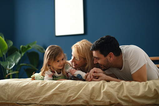 Close up of a couple with their baby girl lying on their bed facing down.  They are looking at each other and smiling while being affectionate.