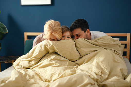 Little girl is sitting on the bed between her parents and together they are hiding behind the covers.