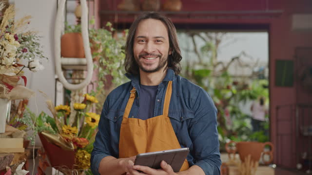 Portrait of a male employee inside commercial shop wearing apron and holding tablet device smiling at camera
