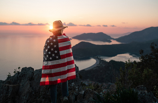 Proud woman covered with the flag of the United States of America standing on the top of the mountain at sunset
