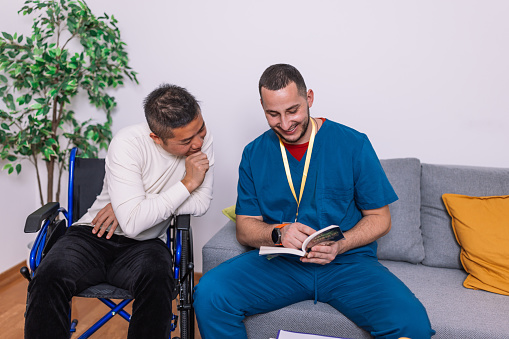 Smiling young Caucasian male doctor reading a book to a mid adult Chinese patient at home