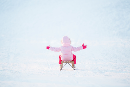 Happy little girl in pink warm clothes with outstretched arms sitting on sledge and sledding down on snow from hill. Enjoying white winter day at park. Back view.