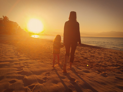 Mom and daughter walk hand in hand along the beach at sunset.