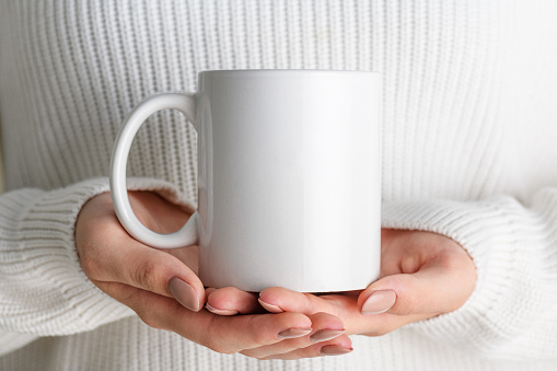 Female hands holding white mug mockup with blank copy space for your advertising text message or promotional content. Girl in white sweater holding white porcelain coffee mug mock up, close up