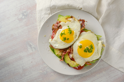 Brunch sandwich with fried egg, avocado and bacon on a white plate and a light wooden table, high angle view from above, copy space, selected focus