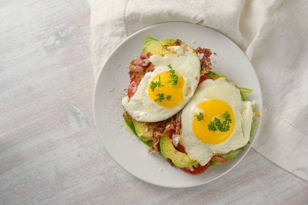 Brunch sandwich with fried egg, avocado and bacon on a white plate and a light wooden table, high angle view from above, copy space stock photo