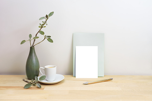 Blank greeting card or invitation mock-up with a pen next to a gray green vase with sage branches and a coffee cup, simple home decor style, copy space, selected focus