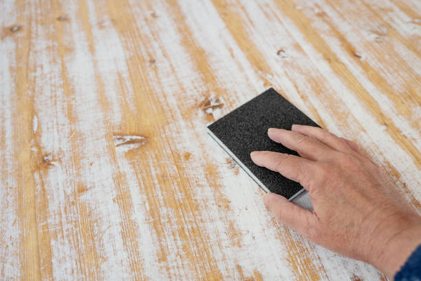 Hand with a sanding sponge removing white paint from the wooden surface of a large table, homemade furniture renovation, copy space, selected focus stock photo