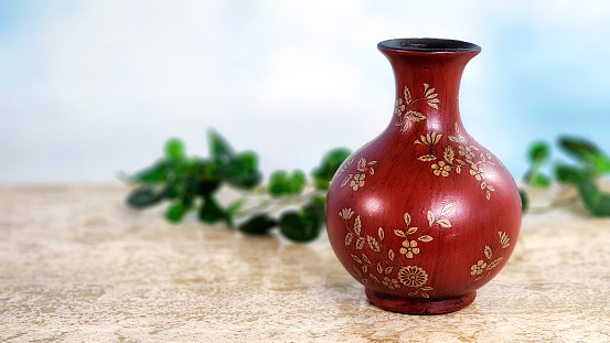 Lovely deep red vase on marble top table by a window.  Greenery behind and lovely sky background.