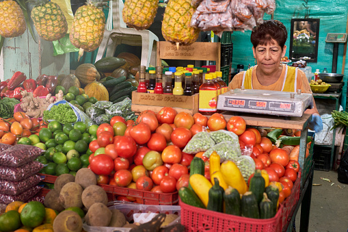 mature woman at market stall selling tomato pineapple and multiple vegetables and fruits next to a food bowl