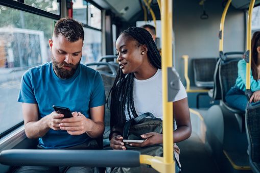 Smiling diverse friends riding together on a bus and using a smartphone. Two multiracial people enjoying travel or ride on public vehicle and having a conversation. Man typing on a mobile phone.
