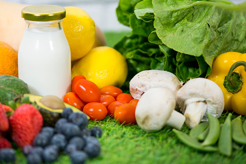 Closeup of a group of healthy fruit, milk, and vegetables sitting on a green grass background. The imperfect, realistic produce consists of mushrooms, peas, lettuce, pepper, blueberries, strawberries, lemons, squash, and avocado.