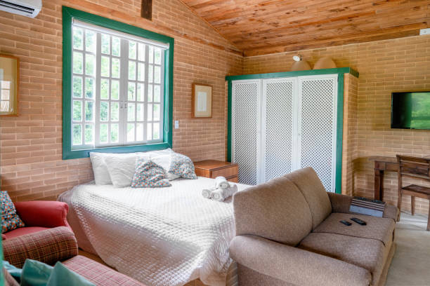 Relaxation room with a wicker bed, white mattress, and pillows. Beige walls with a modern open fireplace on the left. Soft lighting enhances the tranquil ambiance. Bathrobes are neatly placed on the bed, along with a small decorative flower, adding to the spa-like atmosphere. A wooden side table holds wellness accessories, enhancing the room's soothing environment. The setting suggests a focus on comfort and relaxation.