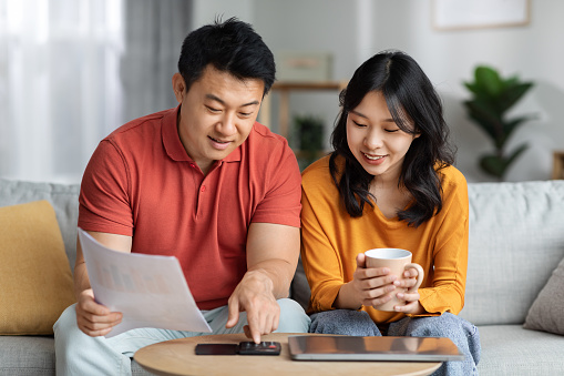 Happy asian family middle aged husband and young wife sitting on couch, holding documents, calculating monthly spending, tracking expenses at home, satisfied with result, family budget concept