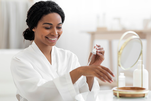 Smiling beautiful black woman applying anti-aging serum on her hand at home, attractive african american female in white silk robe sitting in front of mirror, making beauty routine after bath