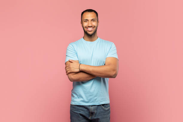 uomo fiducioso. ritratto di ragazzo afroamericano felice in piedi con le braccia conserte e sorridente, in posa su priorità bassa rosa - t shirt shirt pink blank foto e immagini stock