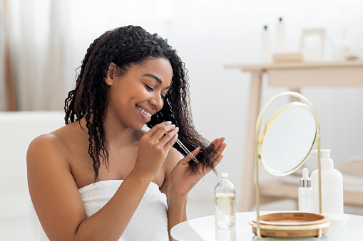 Happy Black Lady Applying Moisturising Oil On Damaged Hair Ends After Shower, Smiling African American Woman Sitting Near Mirror In Bathroom Interior, Making Haircare Routine At Home, Closeup