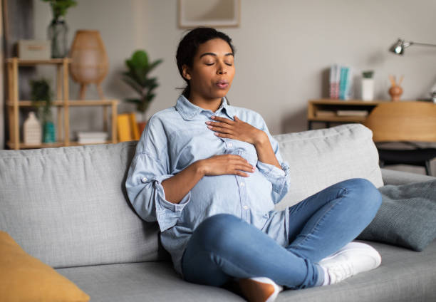 Pregnant African American Woman Doing Breathing Exercise Sitting At Home Pregnancy Yoga. Pregnant African American Woman Doing Breathing Exercise With Eyes Closed Relaxing Sitting On Couch At Home. Relaxation And Childbirth Healthcare Concept muscular contraction stock pictures, royalty-free photos & images