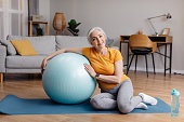 Happy aged woman resting on yoga mat with fitness ball, smiling at camera while taking break from her domestic training