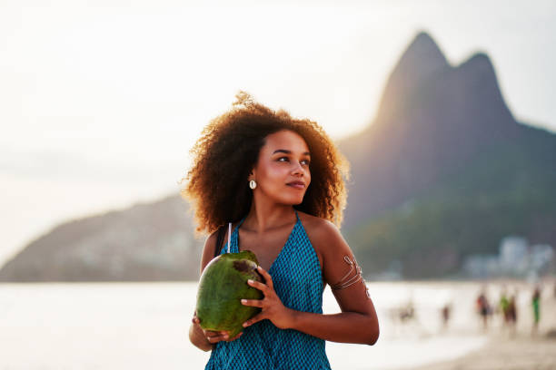 waist up portrait cheerful young brazilian afro hairstyle woman walking on the beach holding a coconut water in Ipanema stock photo