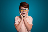 Front view of a boy wearing eyeglasses with his hands on his face looking at camera with amazing expression on blue background. Little amazed boy.