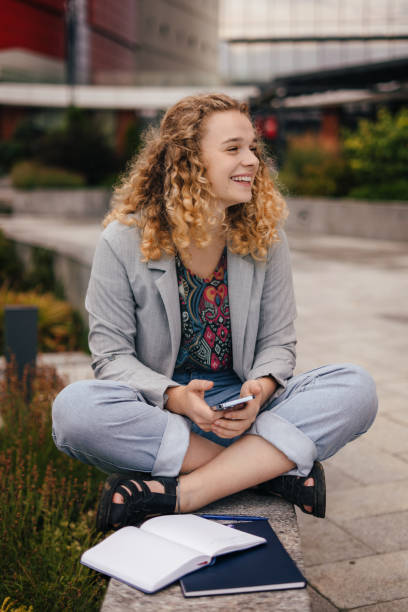 smiling curly haired woman having opened notepad, using cellphone for surfing internet and chatting in online chat, sitting outdoors against blurred background. happy lifestyle. sitting woman outdoors. mobile phone communication. people lifestyle. - surfing wireless vertical outdoors lifestyles imagens e fotografias de stock