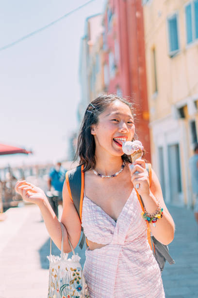 Portrait of a Cheerful Chinese Asian Tourist Young Woman Licking Sweet Italian Gelato Ice Cream while Visiting Venice Italy on Holiday in the Summer Portrait of a Cheerful Chinese Asian Tourist Young Woman Visiting Venice Italy on Holiday during the Hot Summer licking ice cream cone stock pictures, royalty-free photos & images