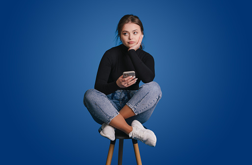 Portrait of attractive minded girl sitting on chair using phone chatting isolated overblue color background