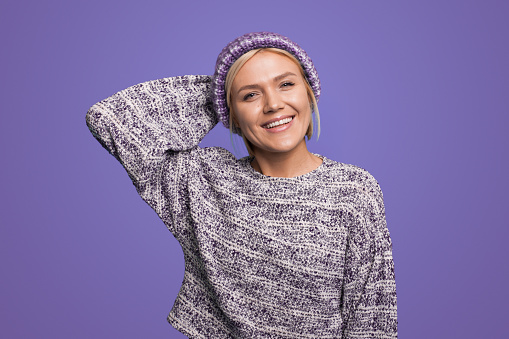 Young beautiful fair-haired woman in knited hat and pullover, smiling looking at camera over purple background.