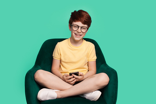 Full body little boy sitting on chair holding in hands mobile cell phone isolated on green background. Lifestyle concept