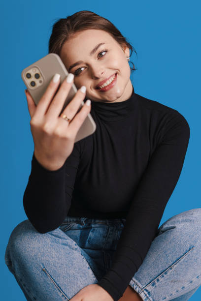 istock Excited woman using mobile phone application sitting on chair isoalted on blue studio background. Texting, communication, cellphone app concept. 1470101119