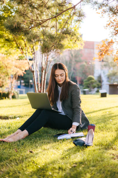 carefree young businesswoman wearing casual clothes using laptop while sitting on grass with disposable cup of coffee. teenager student girl browses internet, typing, working in park outdoors - surfing wireless vertical outdoors lifestyles imagens e fotografias de stock