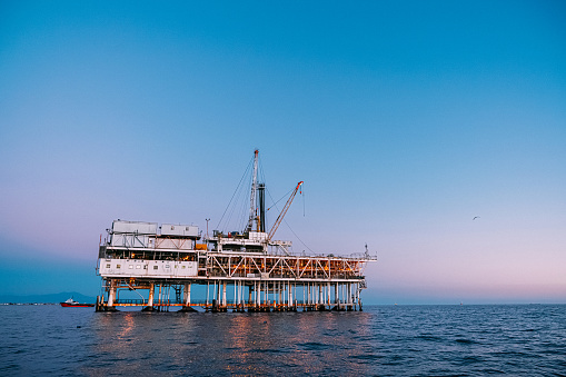 A stunning photograph of an offshore oil rig at dusk off the coast of Huntington Beach, California. The orange, pink, and purple tones of the setting sun highlight the industrial machinery and equipment used in the drilling and extraction of fossil fuels, including crude oil and natural gas. 

This image captures the intersection of the energy industry and the natural beauty of the Pacific Ocean, and speaks to issues of fuel and power generation, energy crises, and environmental concerns surrounding the oil and gas industry.