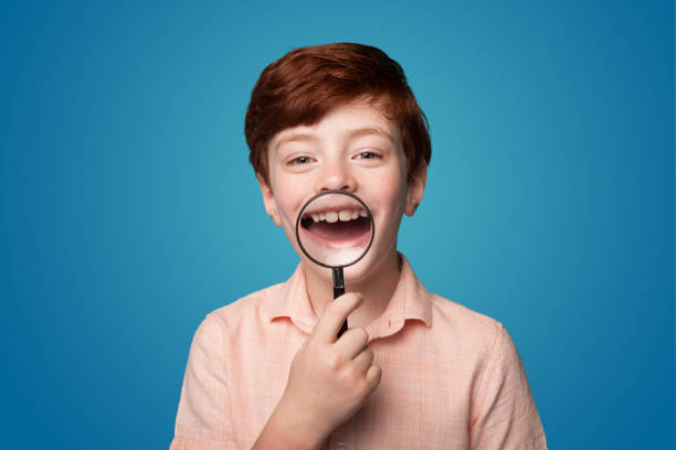 photo of positive candid ginger small boy holding a magnifying glass showing teeth at camera isolated over blue color background - teenager body care adolescence human lips imagens e fotografias de stock