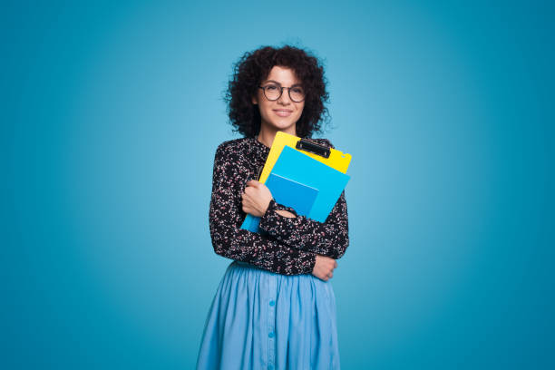 photo de studio d’une femme heureuse posant avec des livres et des cahiers debout sur un fond de studio bleu. grande publicité d’offre éducative - enseignante photos et images de collection