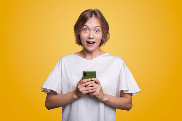 istock Surprised young woman using mobile phone with positive expression looking at camera, standing isolated on yellow background. Happy adorable glad woman rejoices success. 1470101459